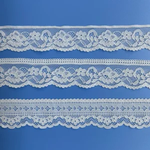 Tres tiras horizontales de encaje elástico blanco con motivos florales sobre un fondo azul liso. Cada tira presenta un intrincado diseño diferente: las dos superiores tienen motivos florales más prominentes y bordes festoneados, y la tira inferior muestra un motivo geométrico y floral más pequeño que se repite.