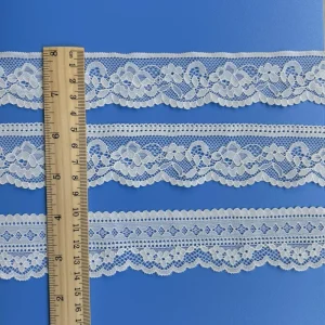 Tres tiras de ribete de encaje floral elástico blanco se colocan horizontalmente sobre un fondo azul. Una regla de madera colocada verticalmente a la izquierda indica la anchura de los adornos. La regla indica que el encaje mide unos 4 cm de ancho. Cada tira presenta delicados motivos florales y geométricos; las dos superiores tienen bordes festoneados y la inferior presenta un diseño más pequeño que se repite.